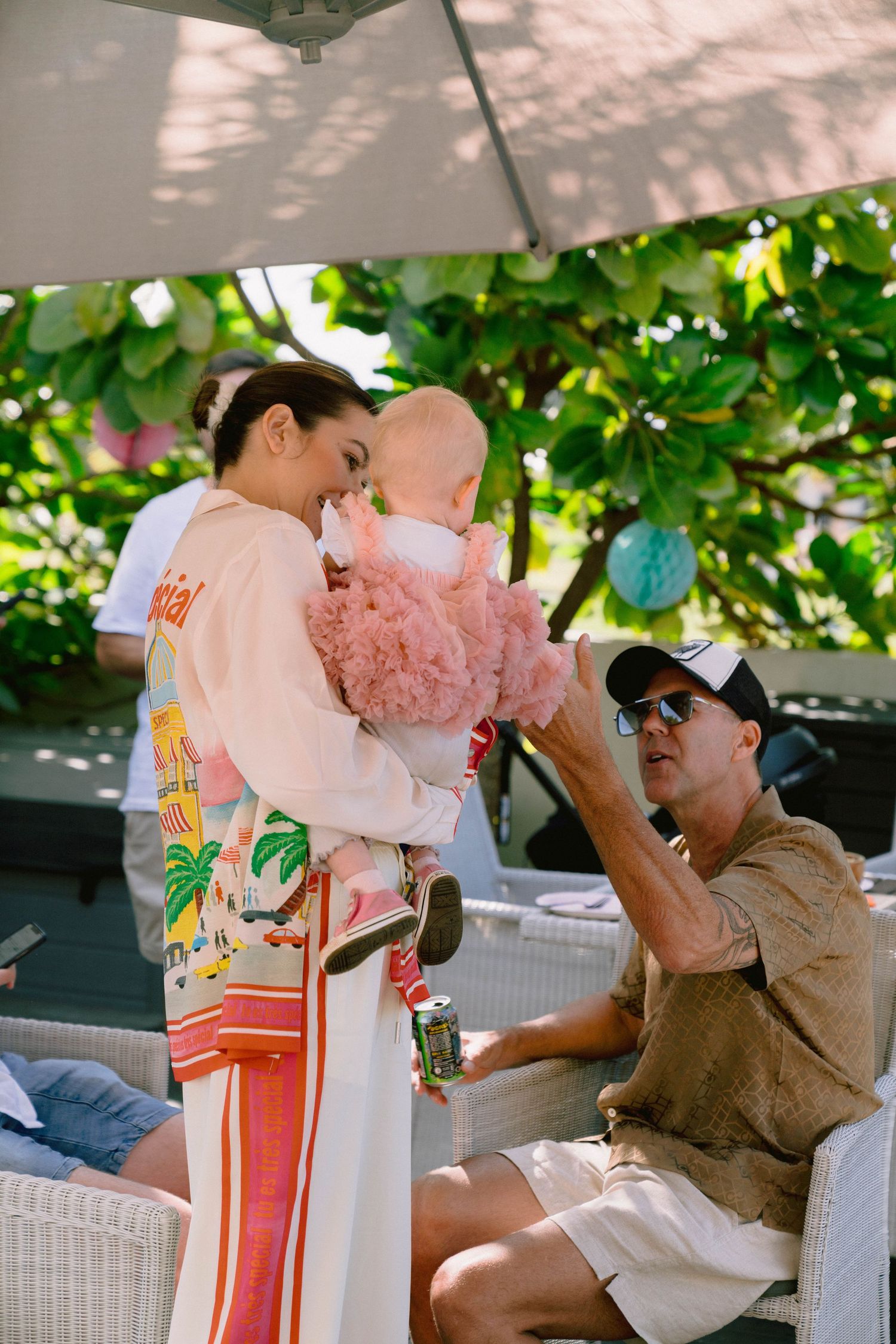 An infant in a pink tulle dress being held at an outdoor gathering beside a pool.