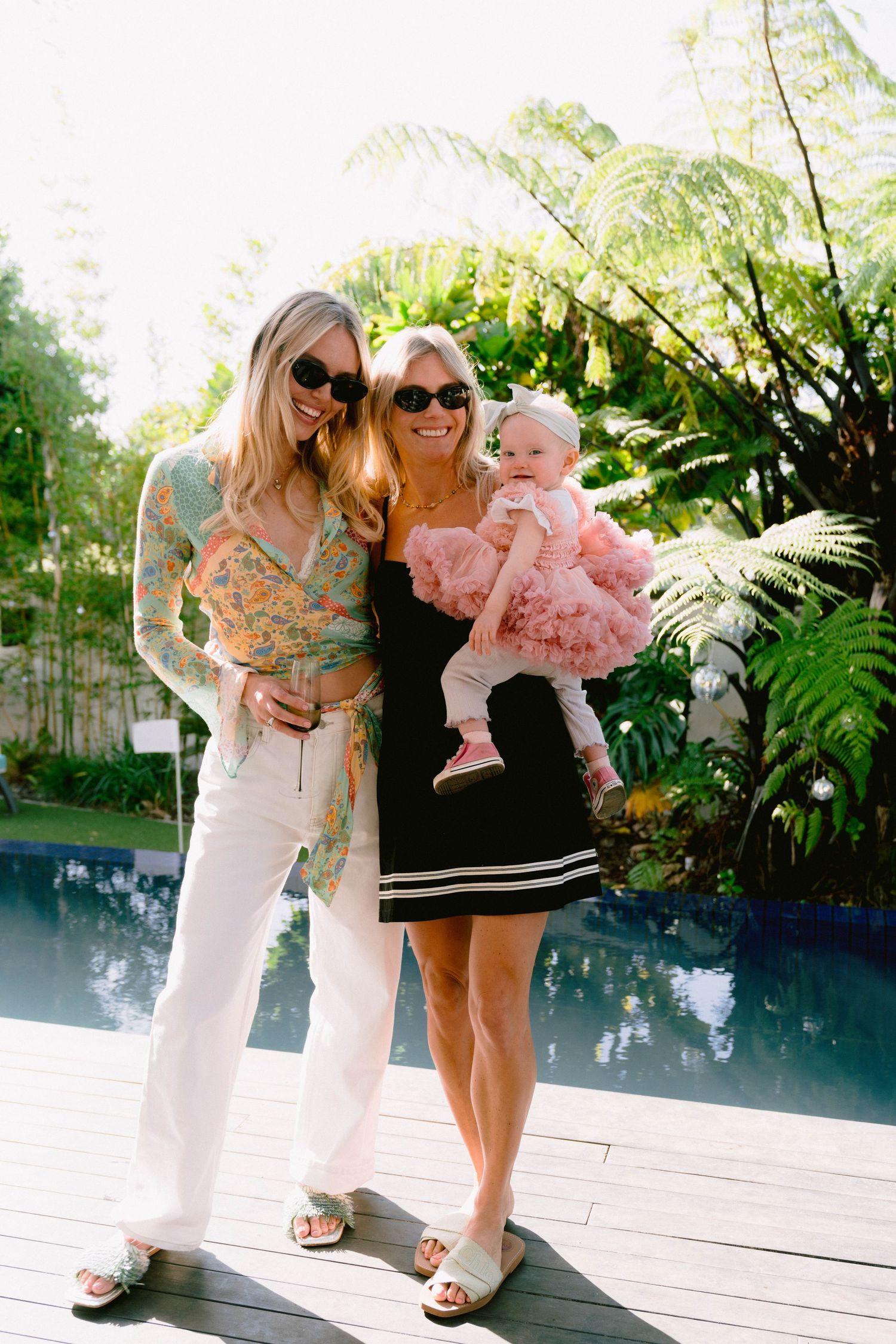Casual outdoor gathering by pool with women and baby in pink dress.