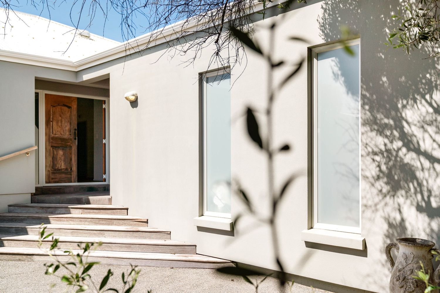 Modern white house exterior with wooden door and concrete steps in dappled sunlight.