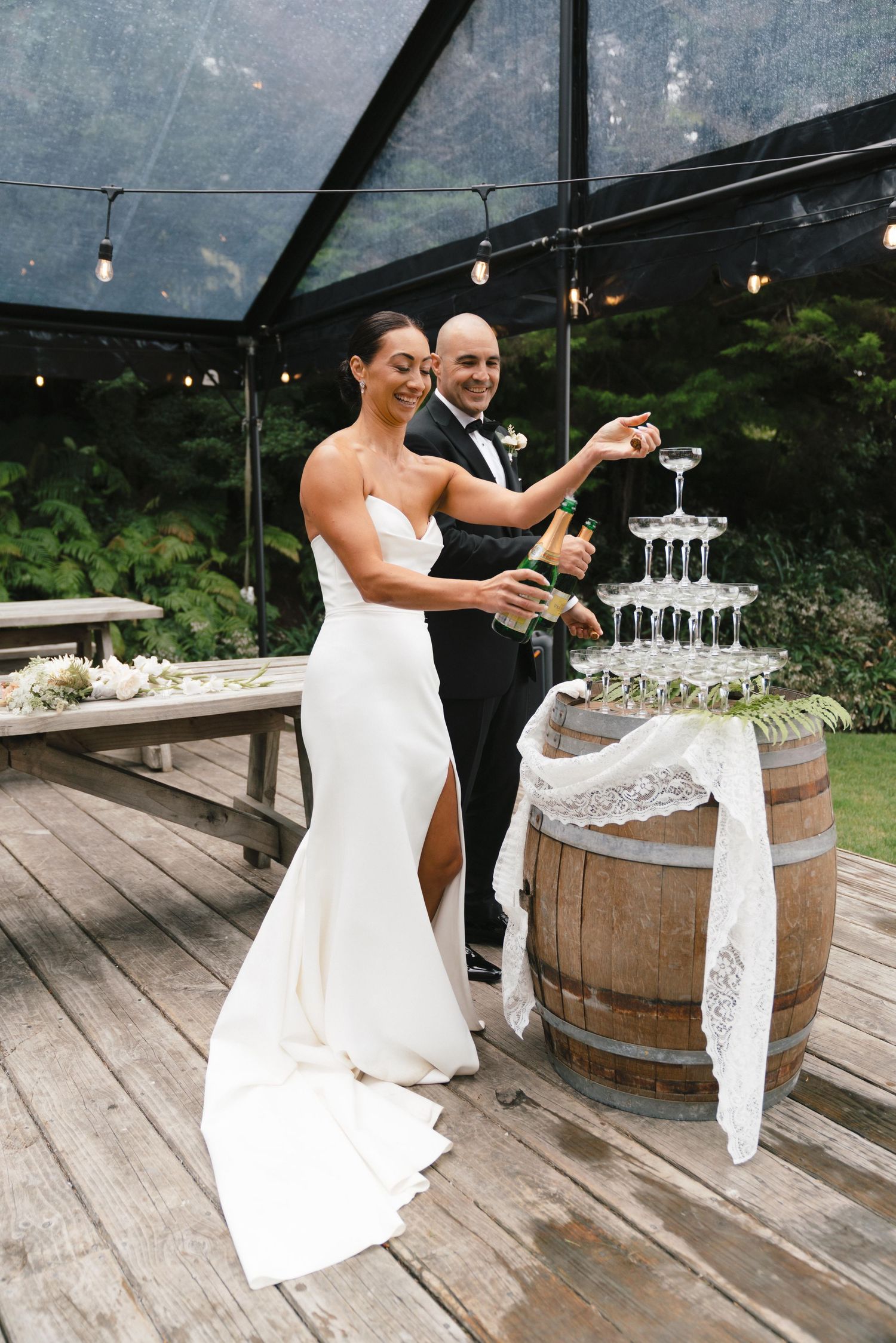 A couple celebrates by popping champagne together on a wooden deck at their outdoor wedding celebration.