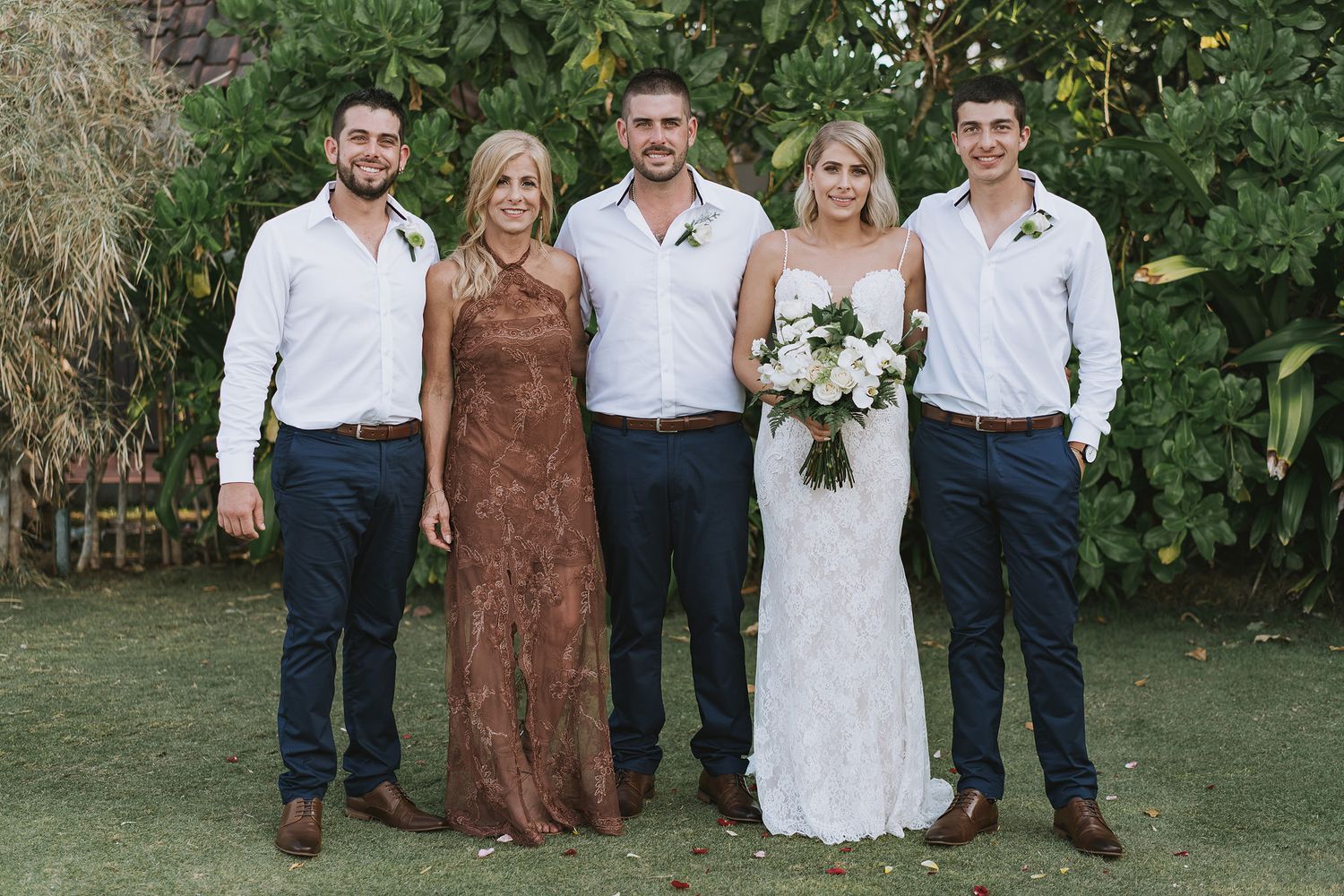 Wedding party poses together on grass with tropical foliage backdrop.