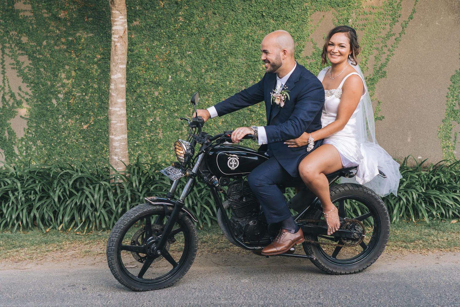 Happy newlyweds having fun riding a black electric bike together through a garden path in playful sequence.