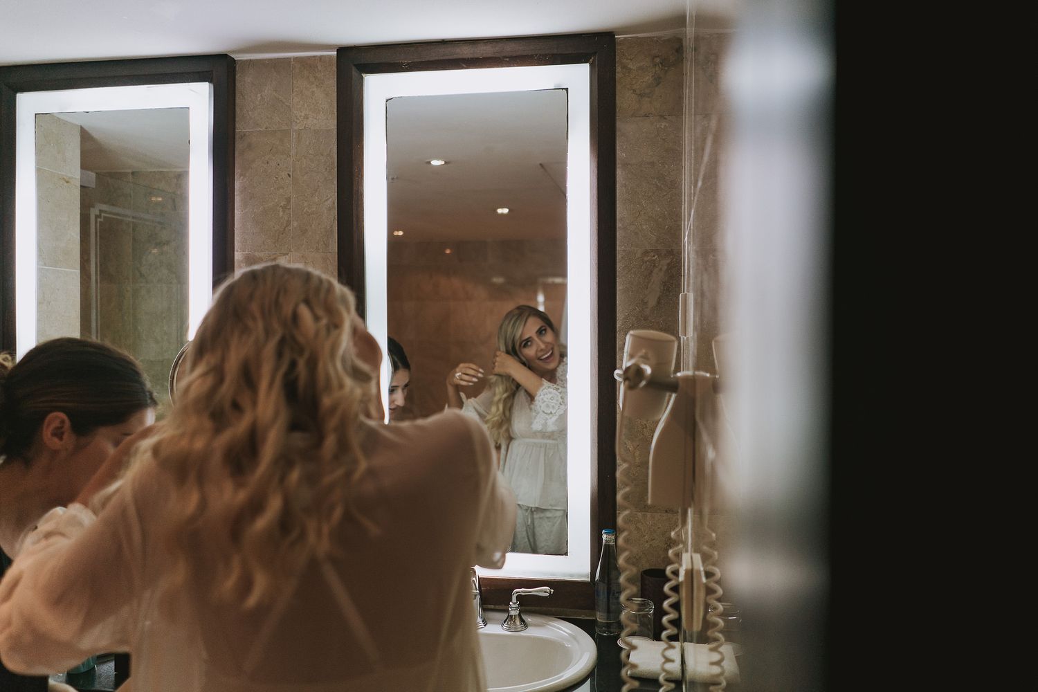 A figure with long wavy hair looks into a well-lit bathroom mirror while getting ready.