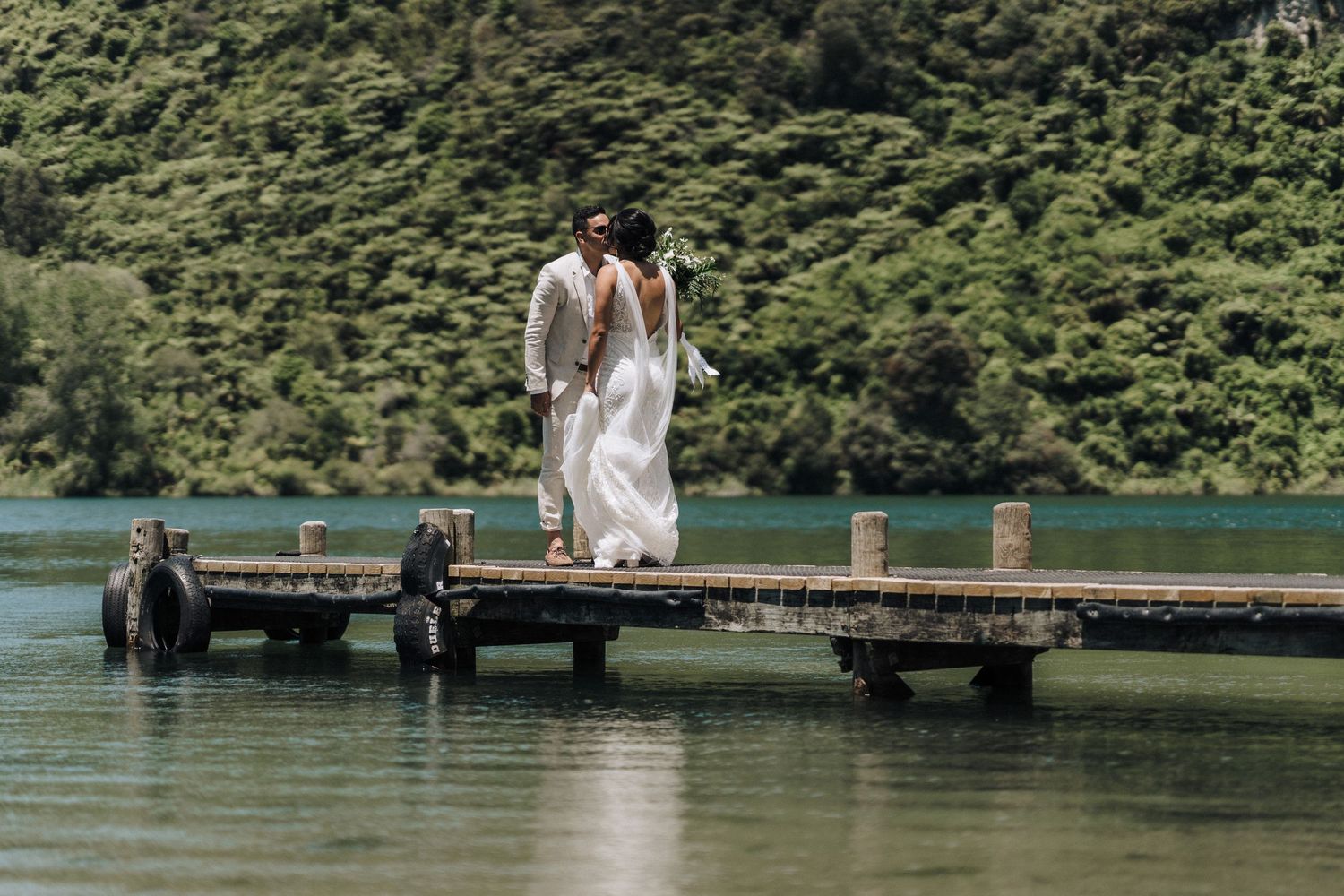 A newlywed couple in white embraces on a wooden dock overlooking a serene lake surrounded by green hills.
