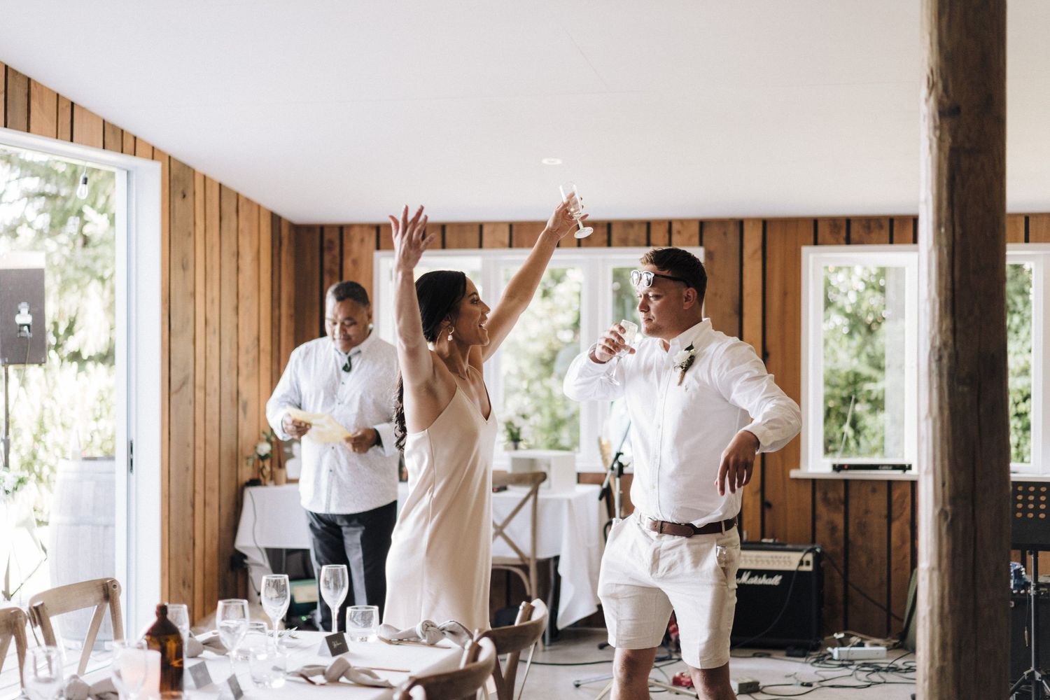 Wedding guests celebrate with raised arms in a rustic wood-paneled reception venue.