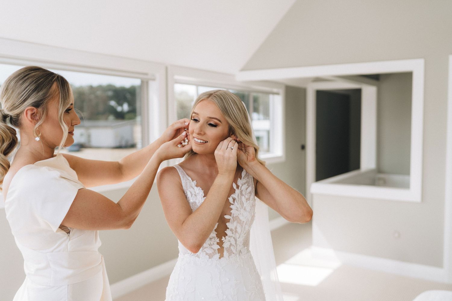 Two women in white dresses share an emotional moment while getting ready in a bright sunlit room.