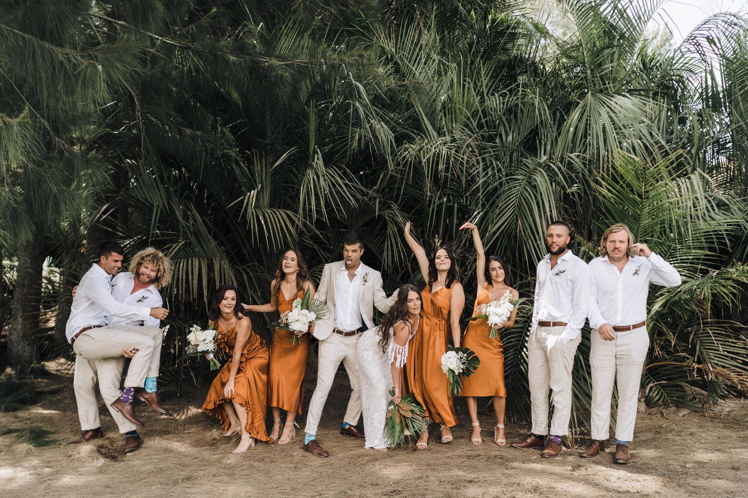 Wedding group photo with palm trees featuring guests in white and burnt orange attire.