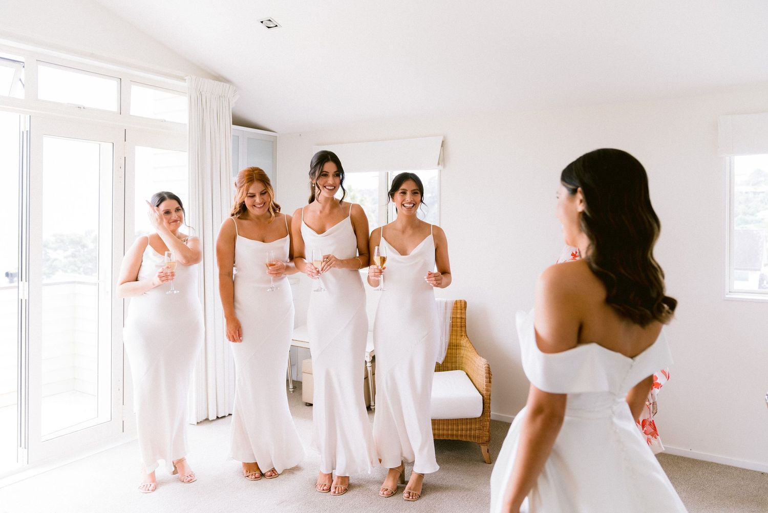 Bridesmaids in white dresses gather in a bright sunlit room during wedding preparations.