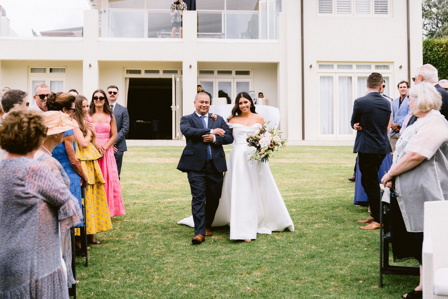 A wedding ceremony processional on a grassy lawn with guests seated in black folding chairs.