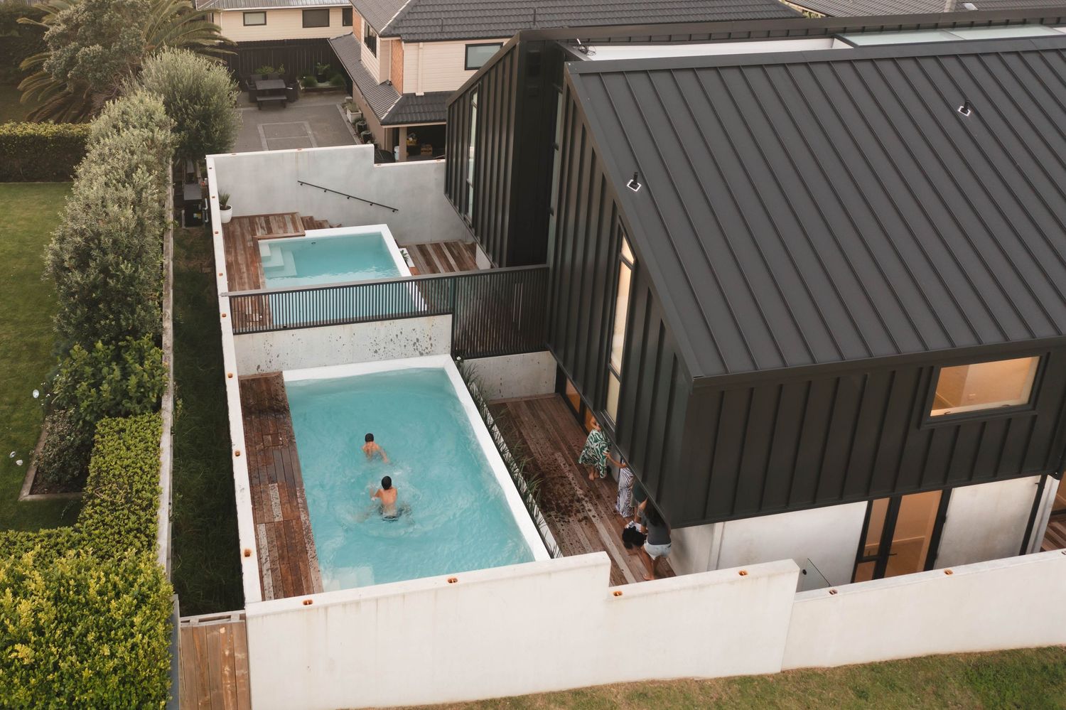 Aerial view of a modern rectangular swimming pool with dark grey house and lush garden landscaping surrounding it.