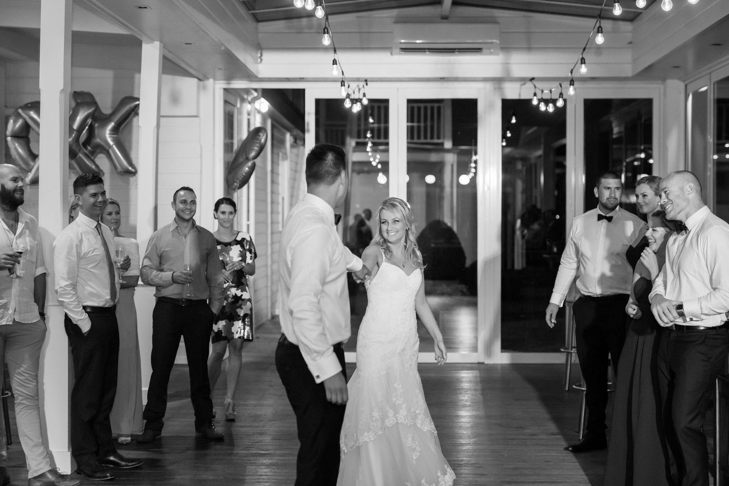 Wedding reception guests dance and socialize in an elegant white-walled venue with dramatic overhead lighting.