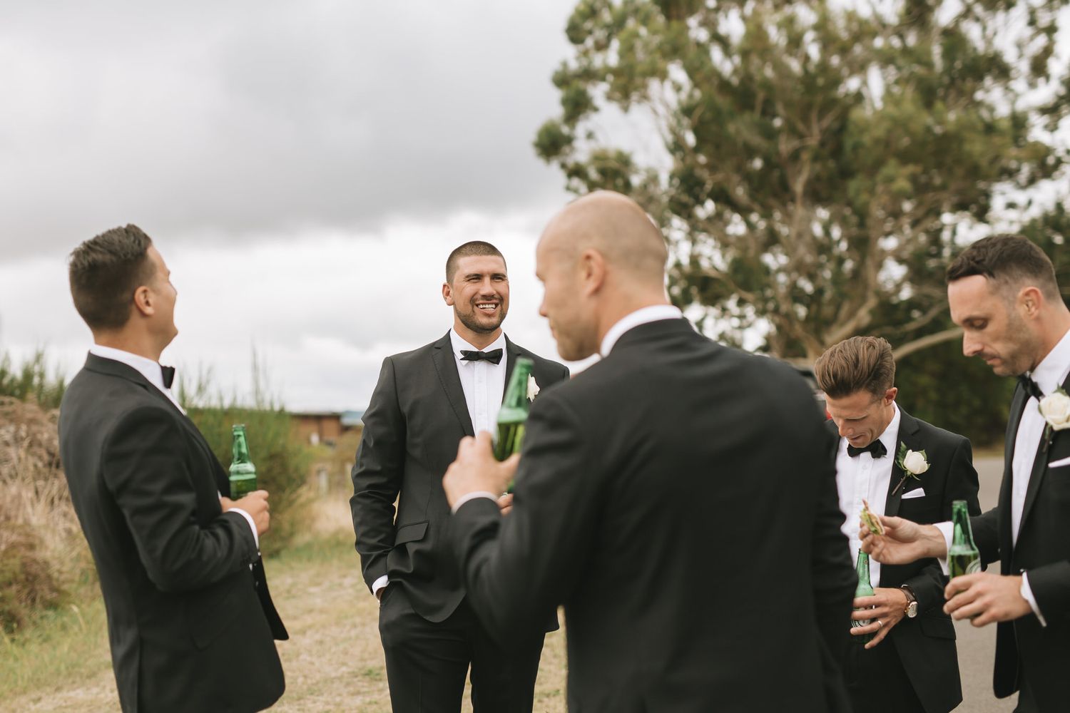 Groomsmen in black suits share drinks and laughs during an outdoor wedding celebration.