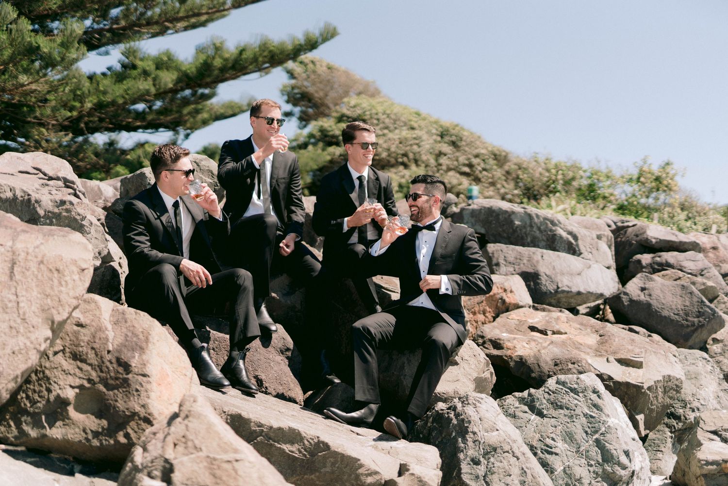A group in black suits sits casually on large rocky boulders against a blue sky on a sunny day.