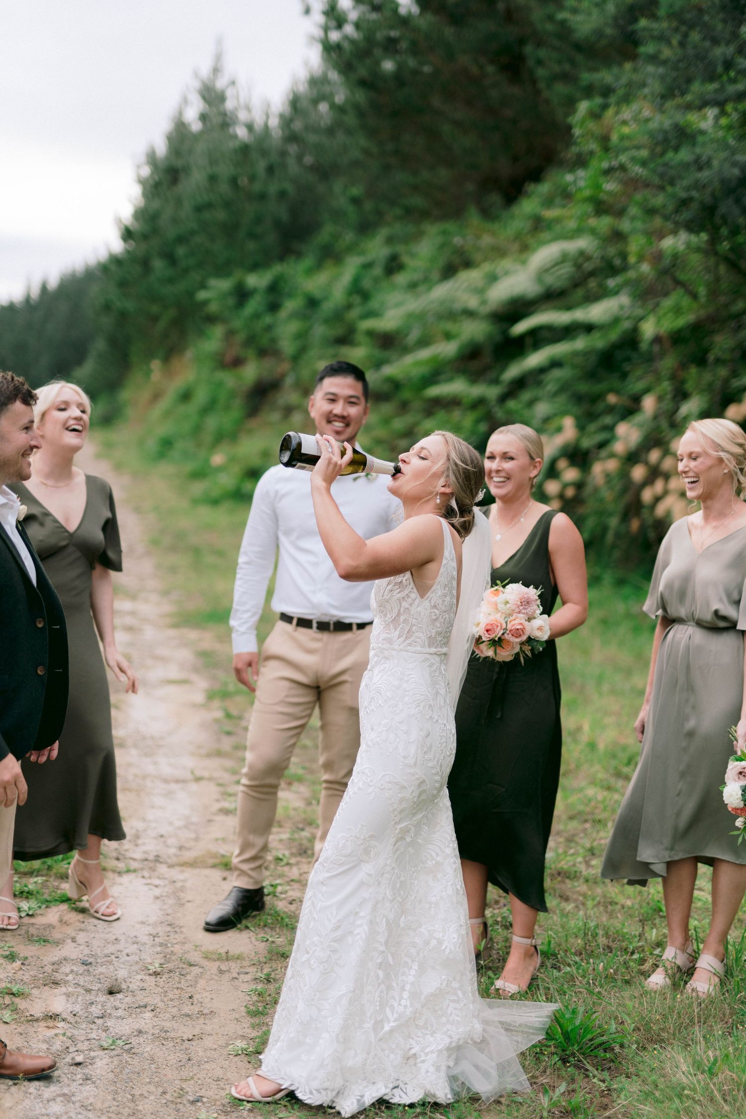 Wedding party shares a laugh while taking celebratory drinks on a scenic forest path.