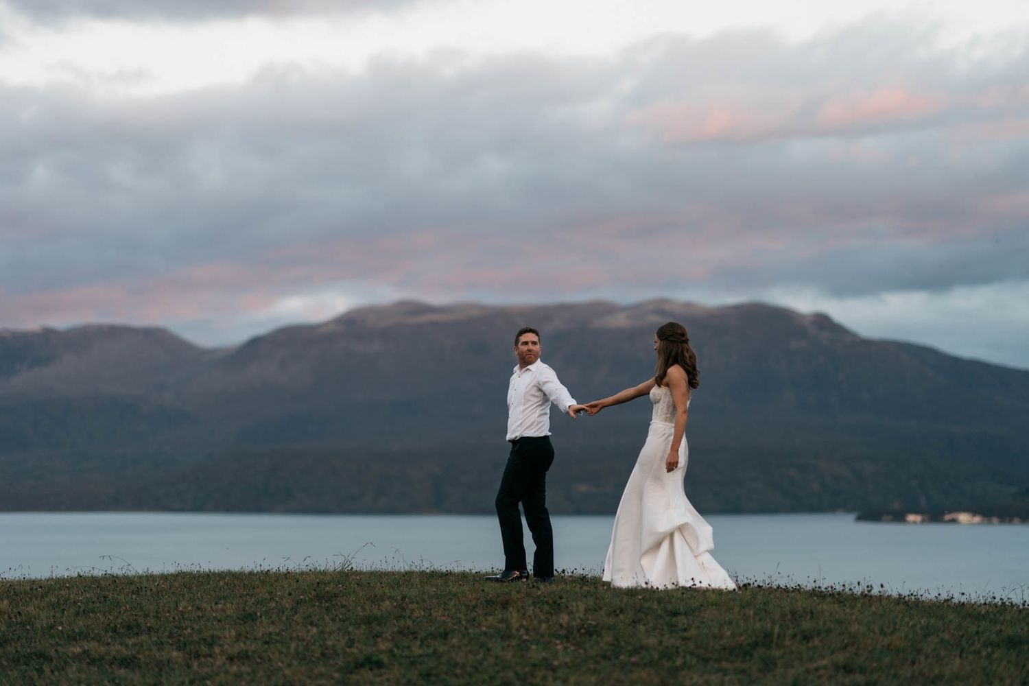 A couple holding hands at sunset overlooking a serene lake surrounded by mountains in moody weather conditions.