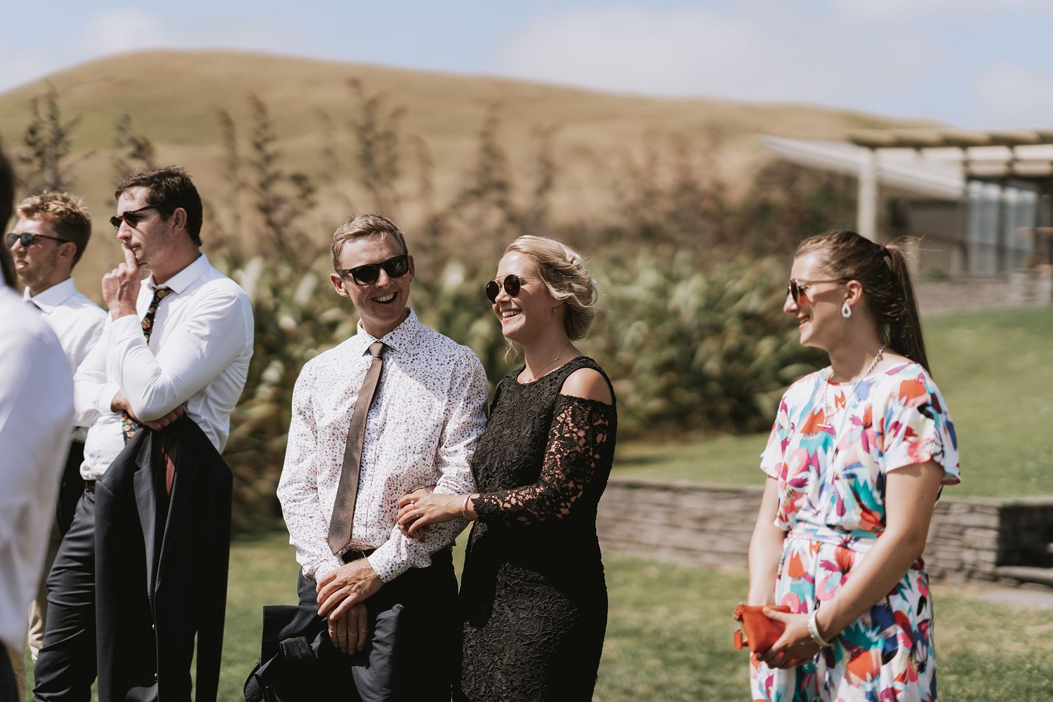 Wedding guests in formal attire stand outdoors against rolling hills on a sunny day.