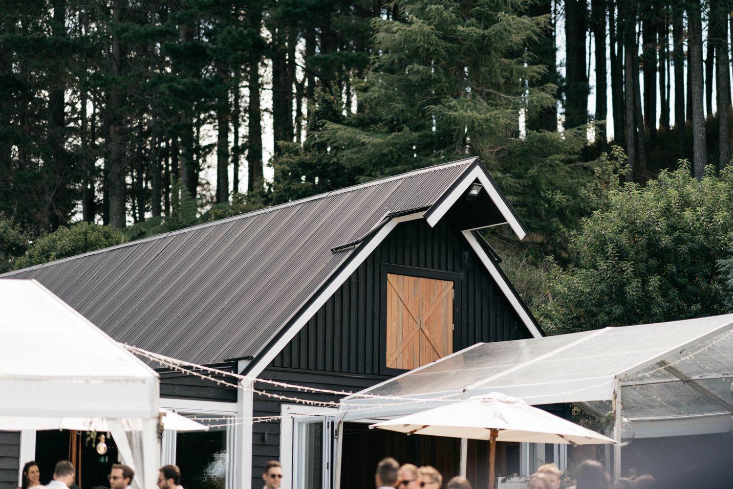Modern black barn-style event venue with metal roof and white reception tents set against a backdrop of tall evergreen trees.
