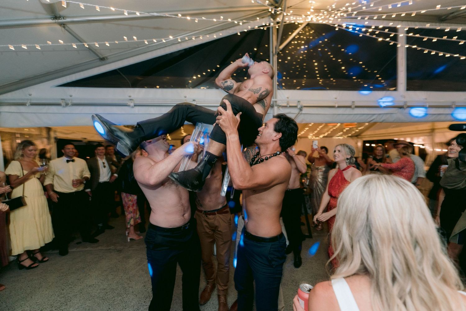 Wedding guests dancing and taking shots together under a large white tent with string lights at night.