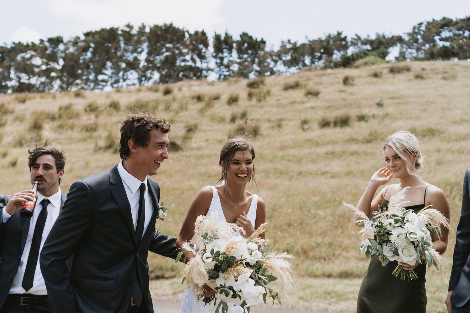 A small wedding party walks together across a scenic grassy hillside on a sunny afternoon, carrying white bouquets.