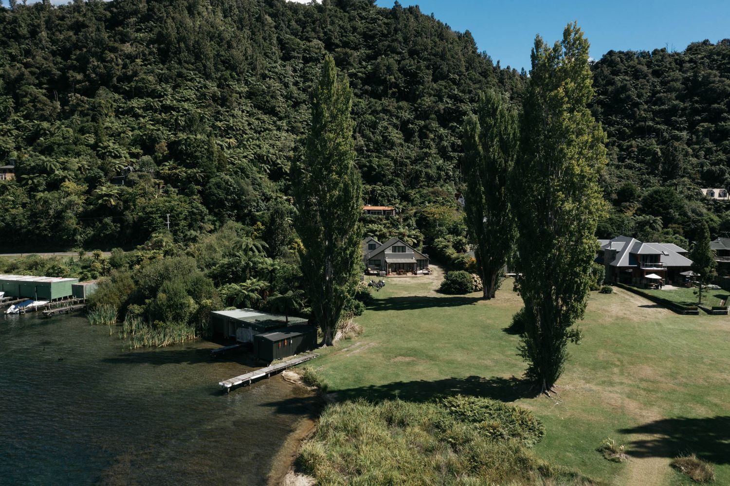 Tall poplar trees line a grassy lakeside camping area with parked RVs and boats against forested hills.