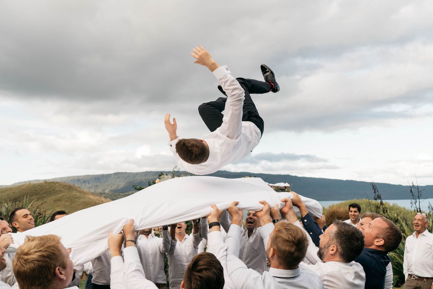 A celebratory group tosses a person wearing white high into the air during an outdoor wedding ceremony against mountain scenery.