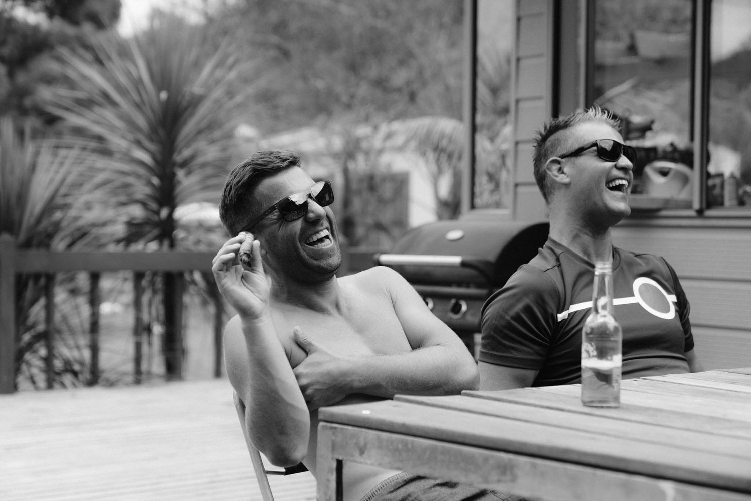 Two people laughing and enjoying drinks while relaxing outdoors at a wooden table on a sunny day.