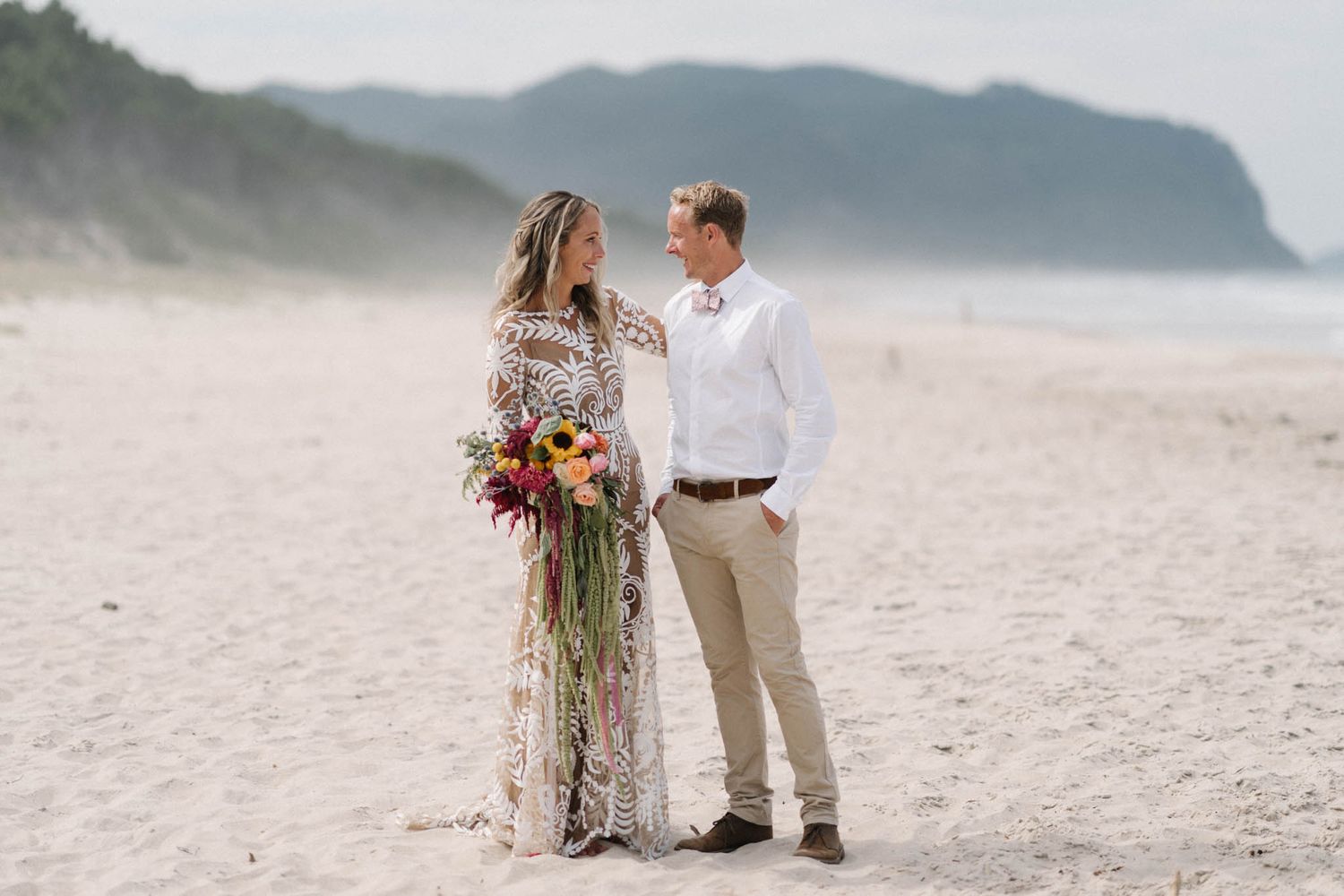 A romantic coastal wedding scene on a misty beach with dramatic cliffs and a colorful wildflower bouquet.