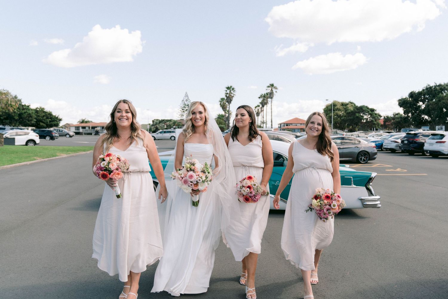 Bridesmaids in light pink dresses walking together in a parking area on a sunny day.