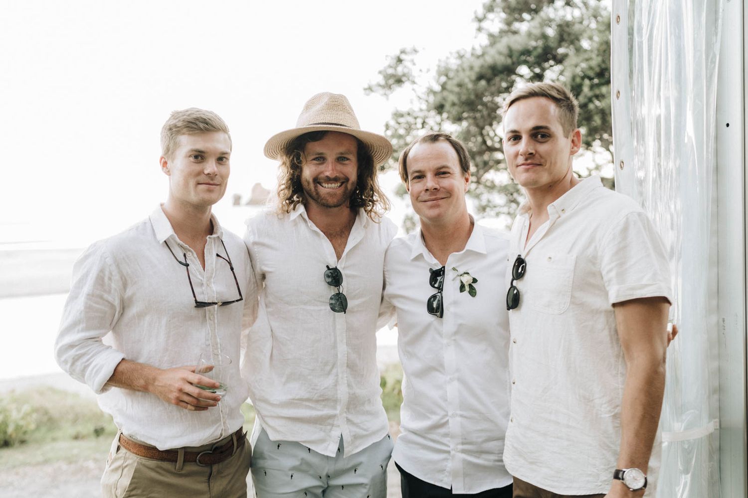 Groomsmen in white shirts and light khaki pants share laughter and drinks at an outdoor wedding celebration.