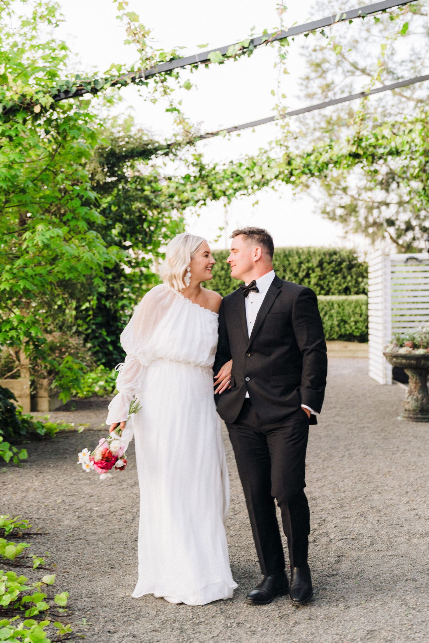Newlyweds walking together under garden trellis with blooming flowers.
