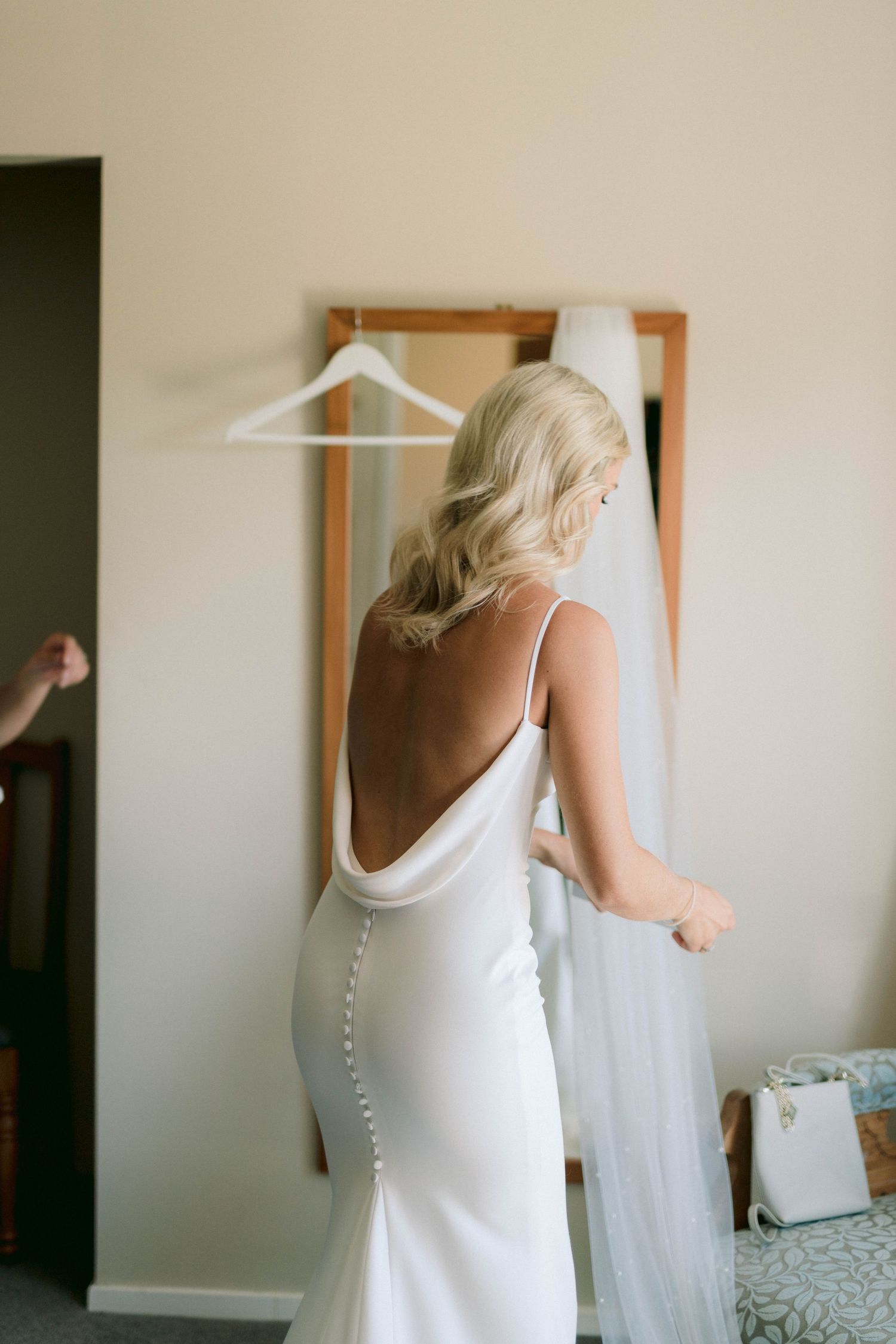 Beautiful backless white wedding dress with delicate button details and cowl neckline captured in natural light.