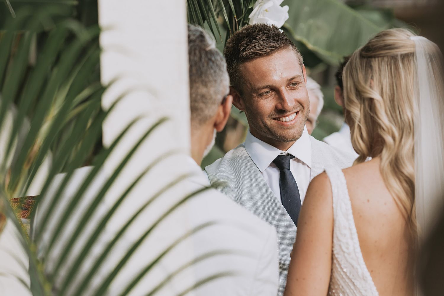 A romantic wedding couple shares intimate moments among tropical palm fronds in soft natural lighting.