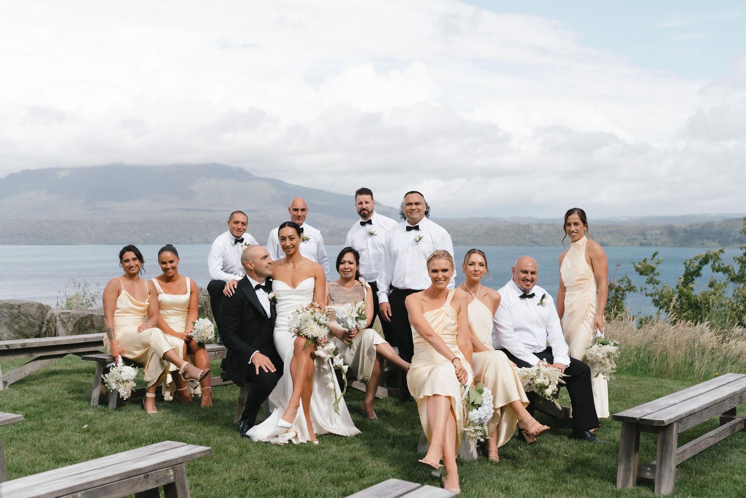 Wedding party in neutral tones poses on wooden bench with scenic coastal backdrop.