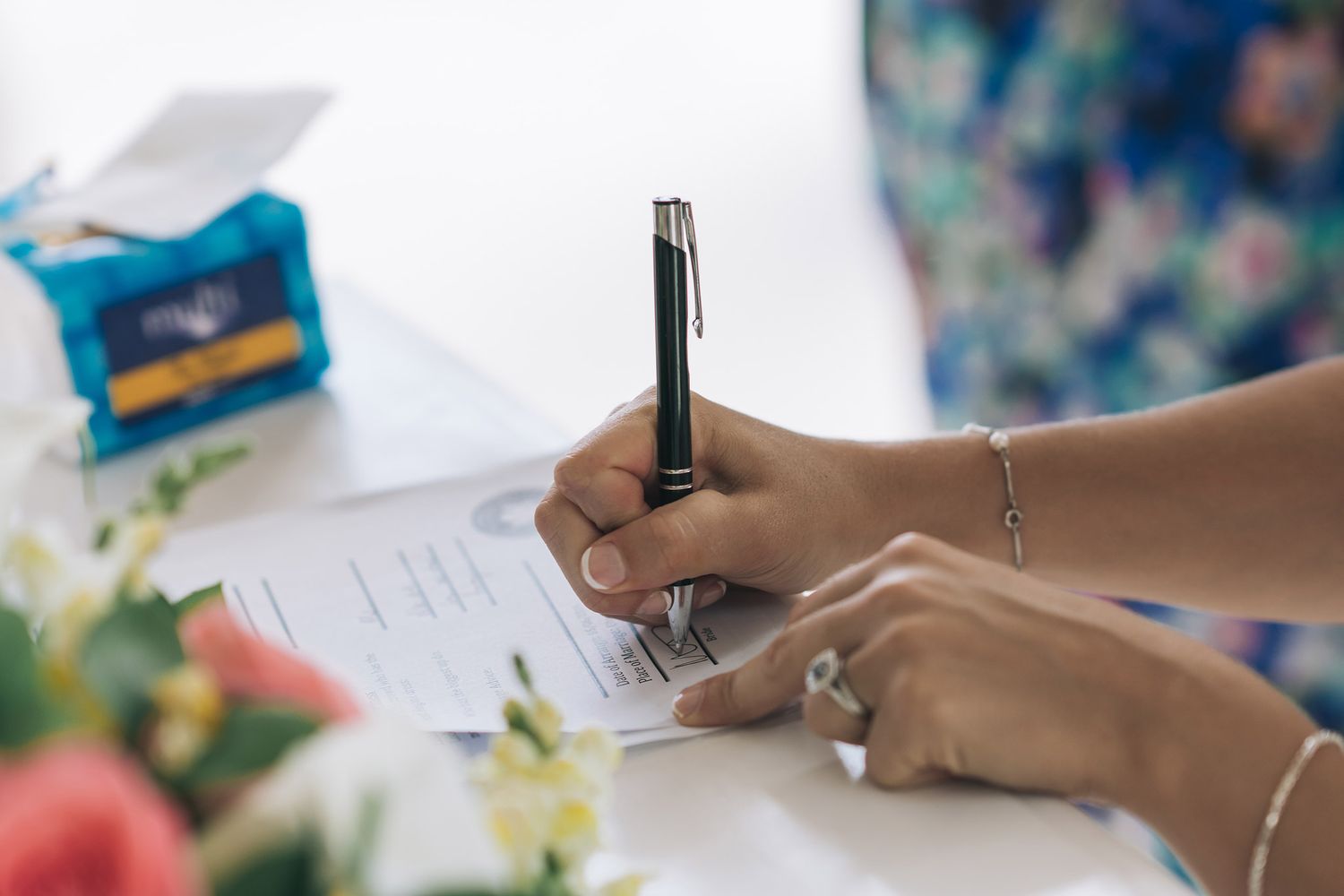 Close-up of hands signing a document with flowers and decorations in the background.