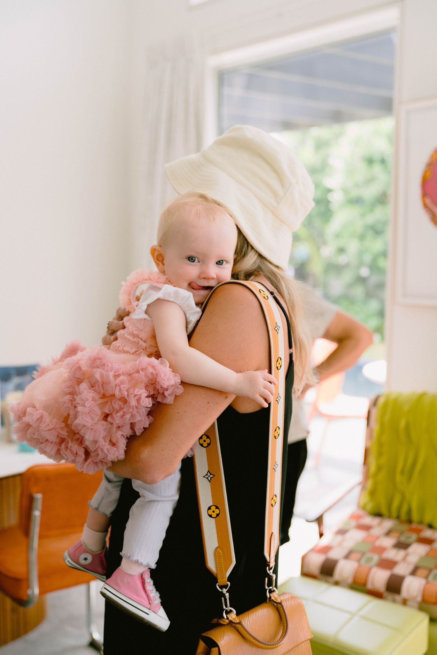An infant wearing a pink tutu dress being lifted playfully in the air in a bright indoor setting.