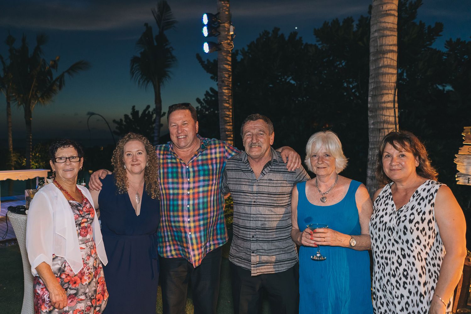 A group poses together at a tropical evening event with palm trees silhouetted against the darkening sky.