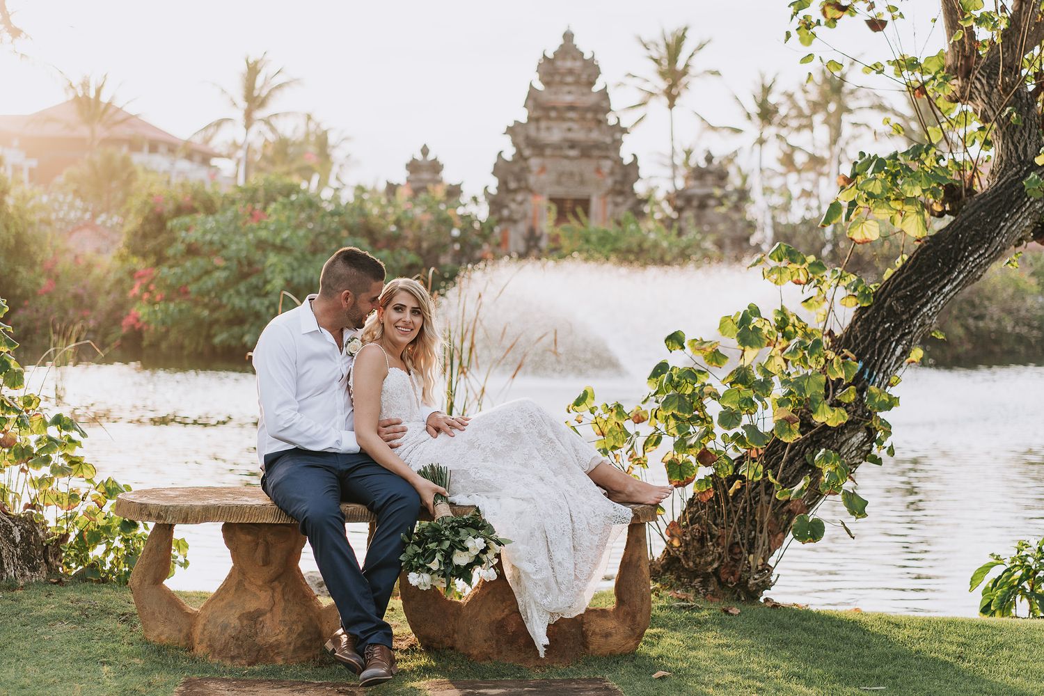 Couple sits on stone bench near traditional Balinese temple during destination wedding.