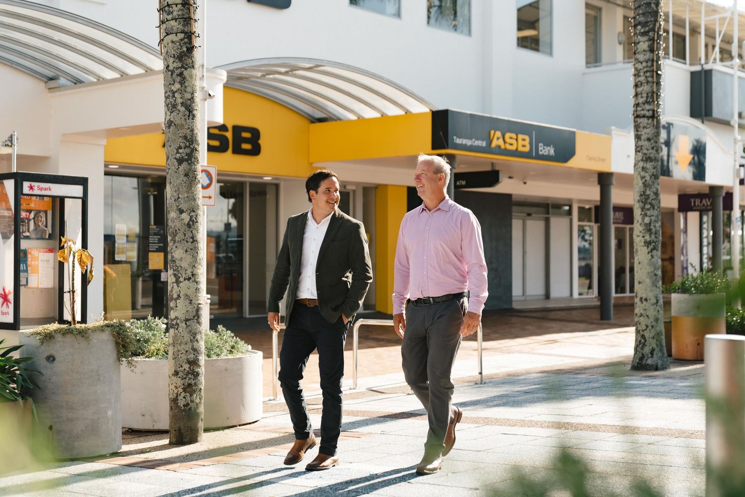 Business partners walking past retail shops with palm trees and modern architecture.