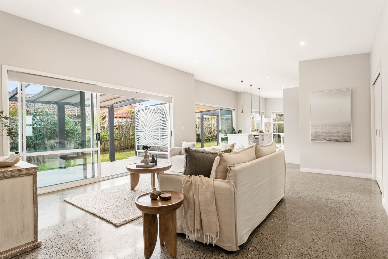 Modern living room with large glass doors, beige sofa, wood side tables and grey carpet in bright neutral tones.