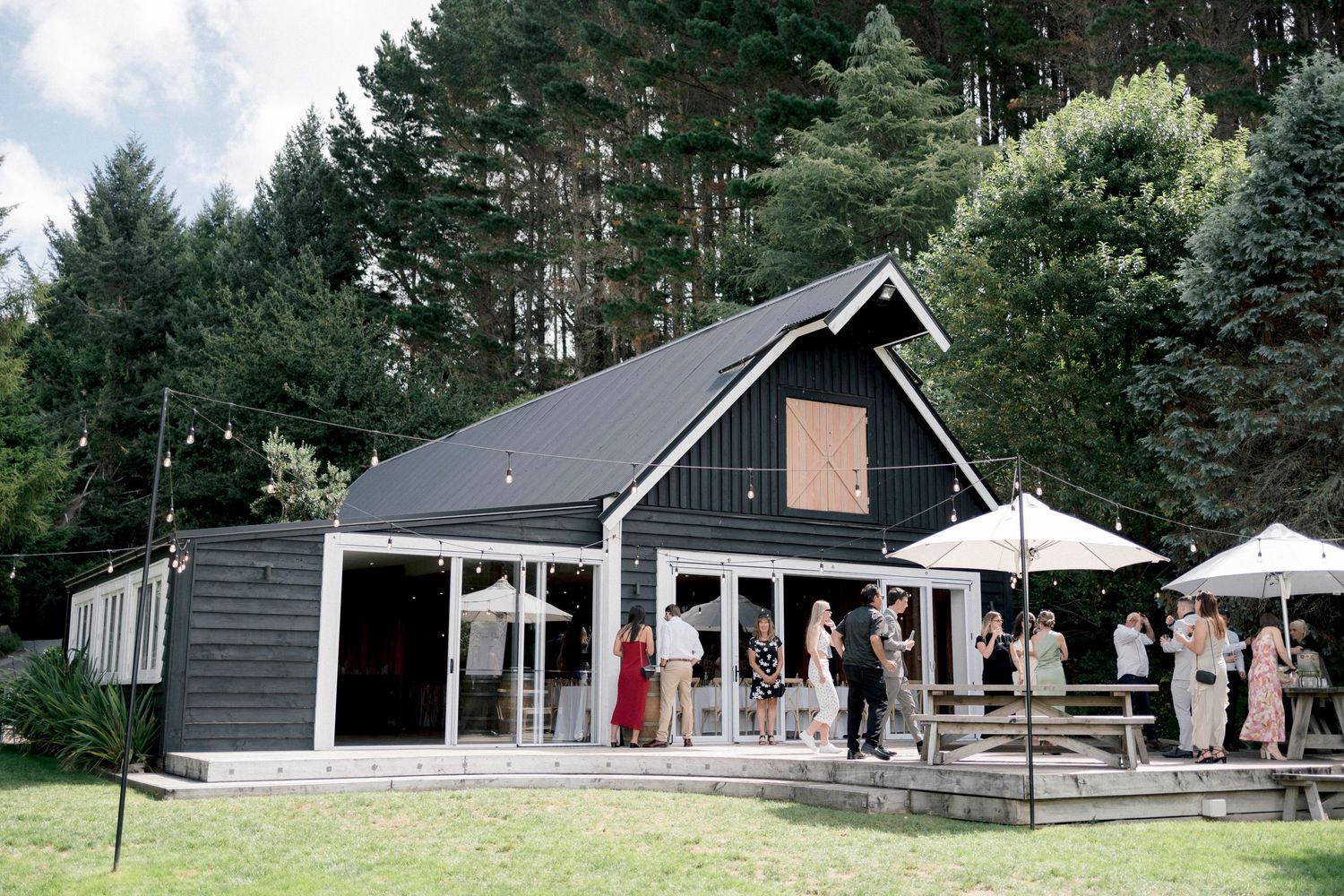 Modern black barn-style wedding venue with outdoor patio area surrounded by tall pine trees.