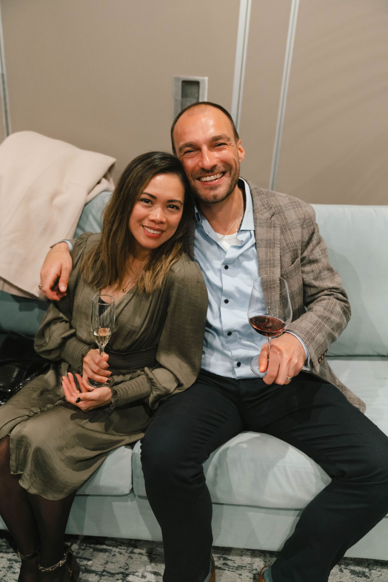 A couple relaxes together on a light-colored couch at an indoor social gathering.