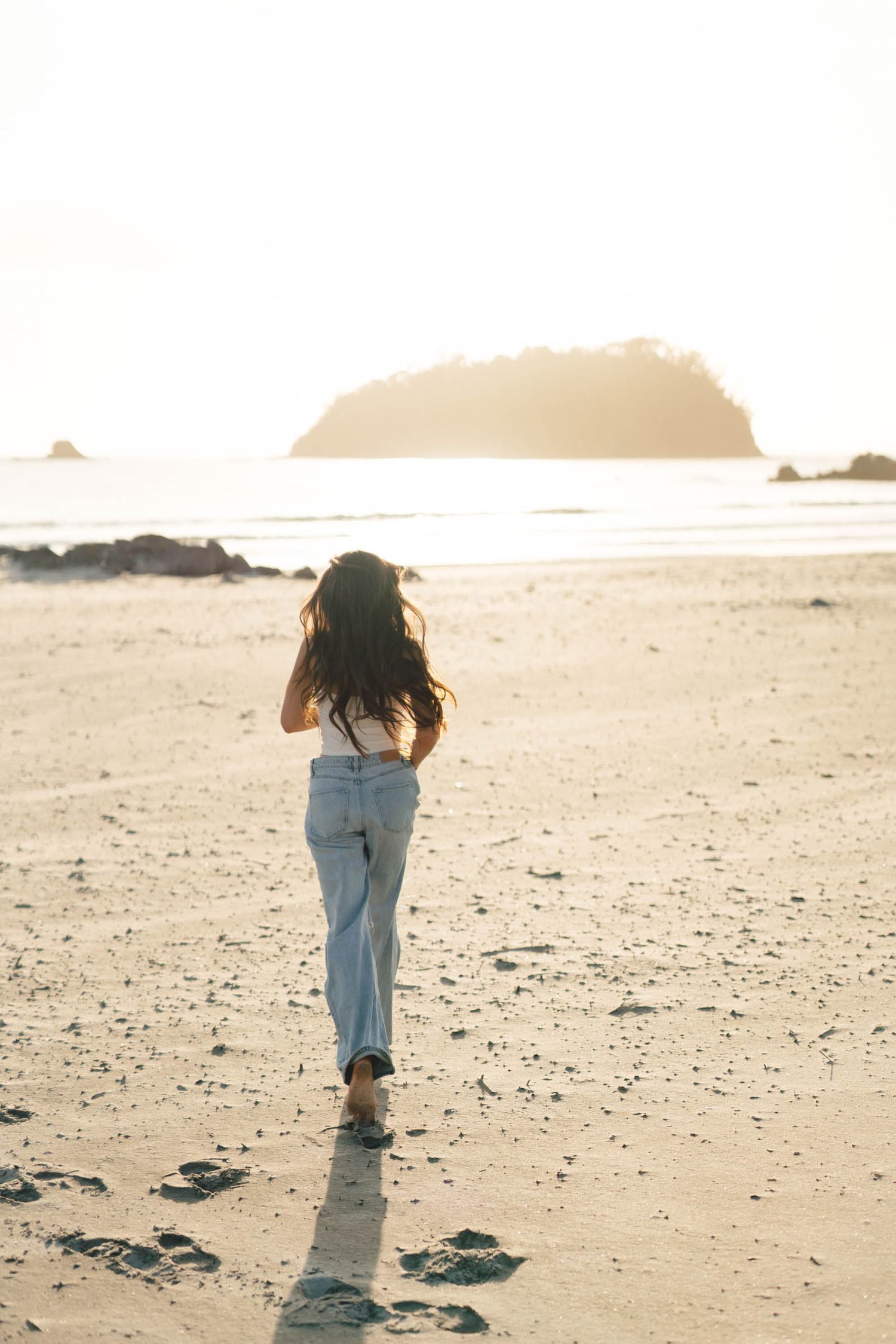 Person walks along empty beach towards distant island during golden hour.