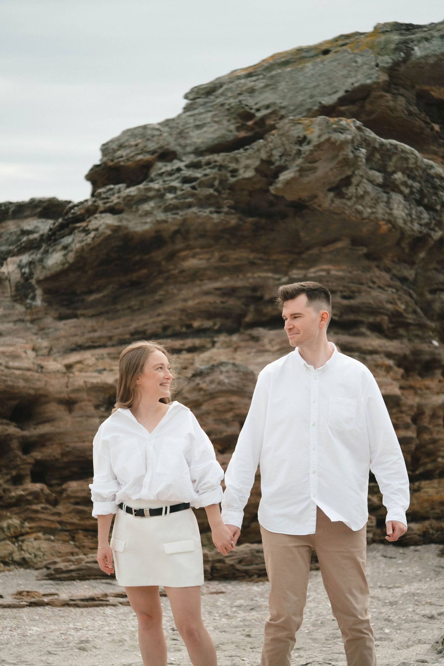 Two people wearing coordinated white outfits laughing together on a beach with rocky cliffs.