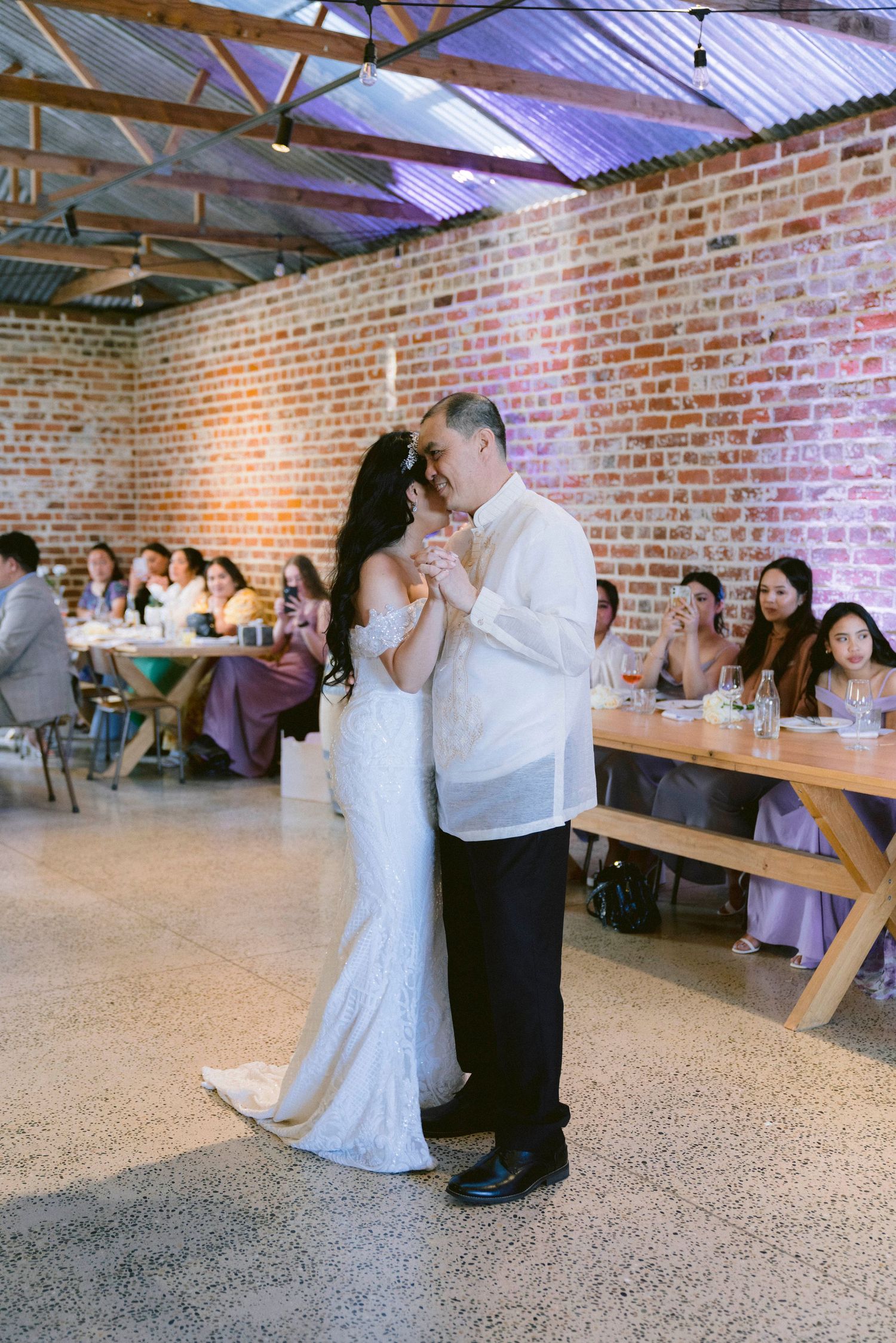 A couple shares their first dance as newlyweds in a rustic brick reception venue with guests looking on.