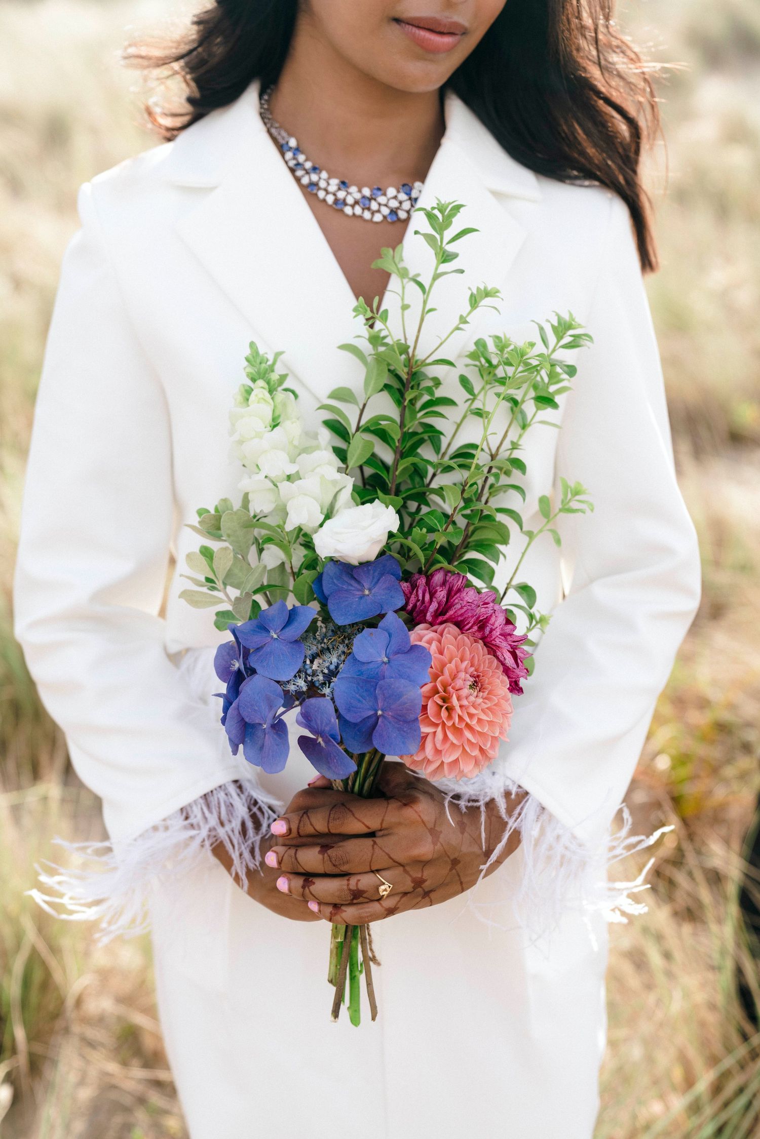 A hand in white holds a vibrant bouquet with deep blue hydrangeas, coral peonies and white flowers.