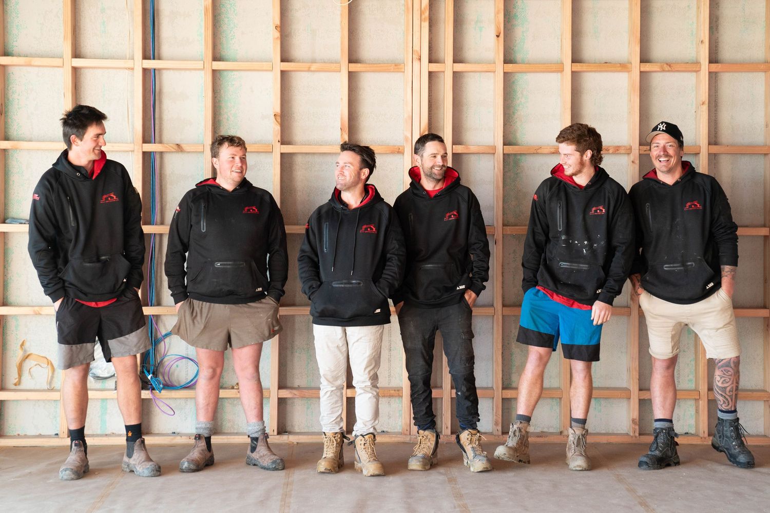 Six construction workers stand casually along a wall wearing casual work attire and boots.