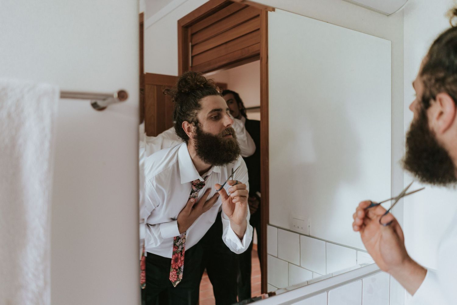 A bearded person in a white shirt and suspenders fixes their bowtie while looking in a bathroom mirror.
