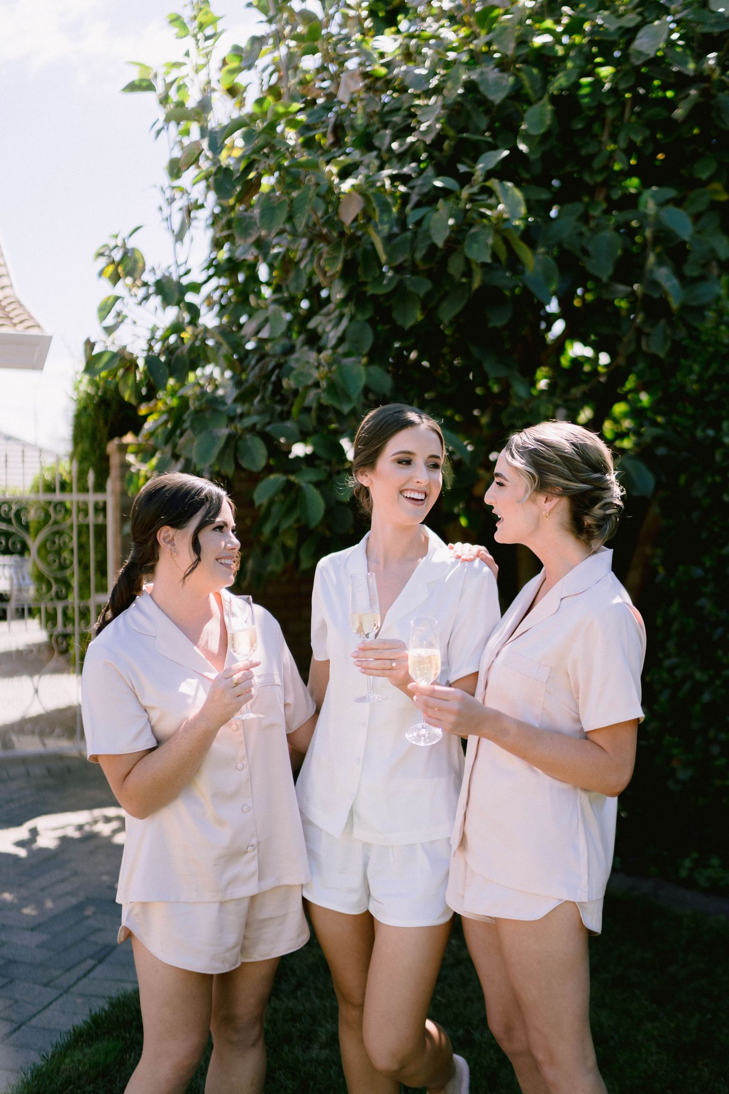 Three women in matching white pajama sets laugh and chat while holding drinks in a sunny garden setting.