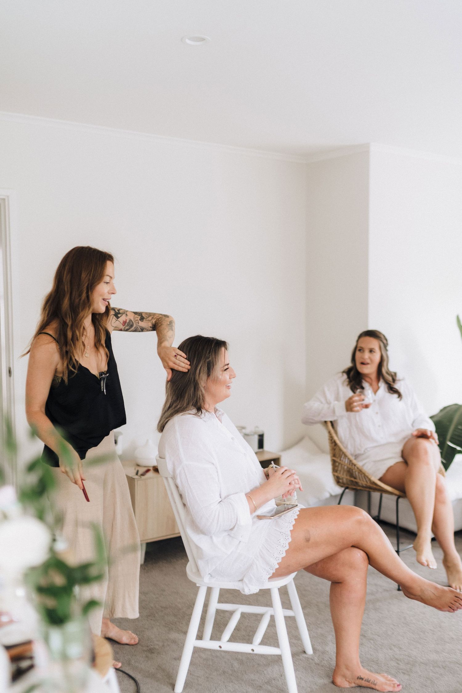 Women getting ready in a bright white room with one seated in a chair while another styles her hair.