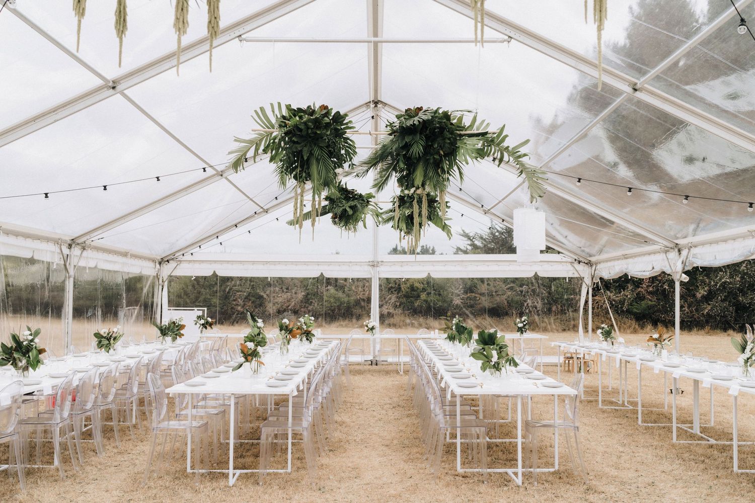 Wedding reception tent setup with white tables and hanging greenery decorations.